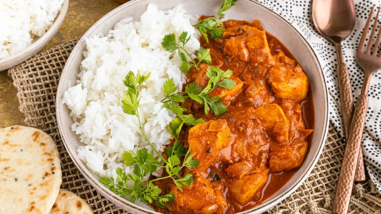 Ein Teller mit dampfendem Reis und cremigem Chicken Tikka Masala, garniert mit frischem Koriander. Dazu Naan-Brot und ein Löffel.