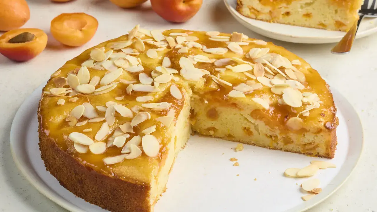 Saftiger aprikosenkuchen mit frischen aprikosen und Mandelblättchen bestreut, ein Stück auf einem Teller daneben.