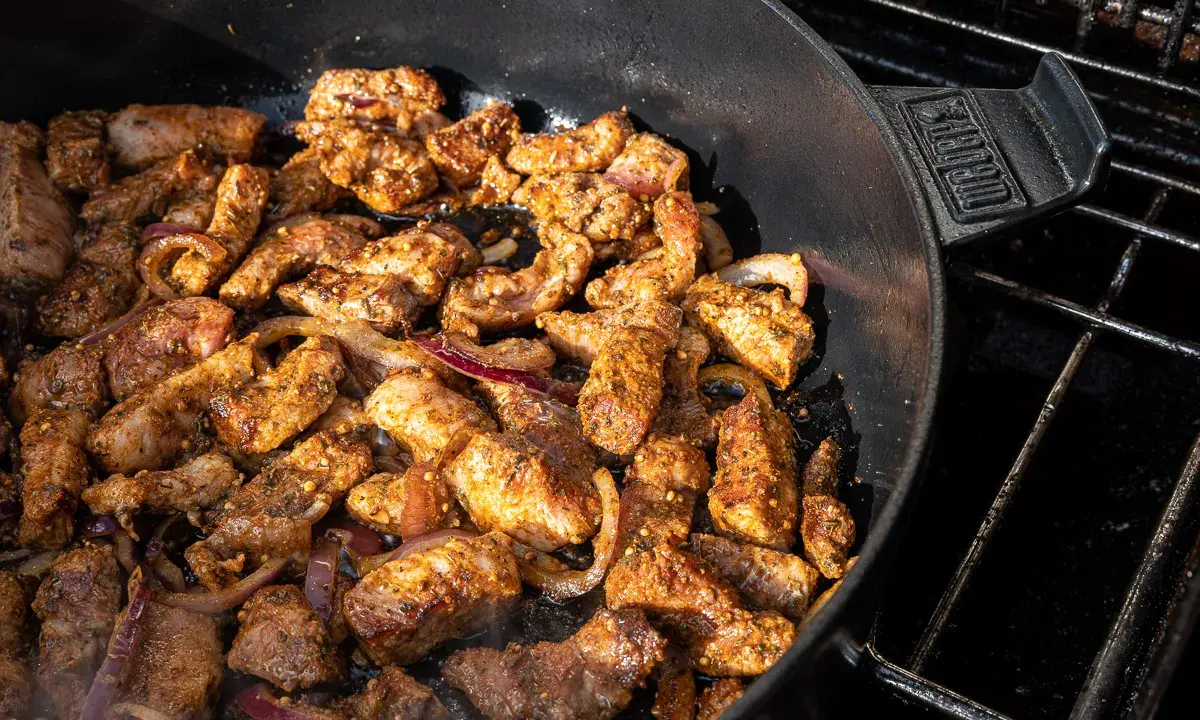 Würziges Gyros brutzelt in einem Dutch Oven auf dem Grill. Zarte Fleischstücke mit Zwiebeln garen in der Pfanne.