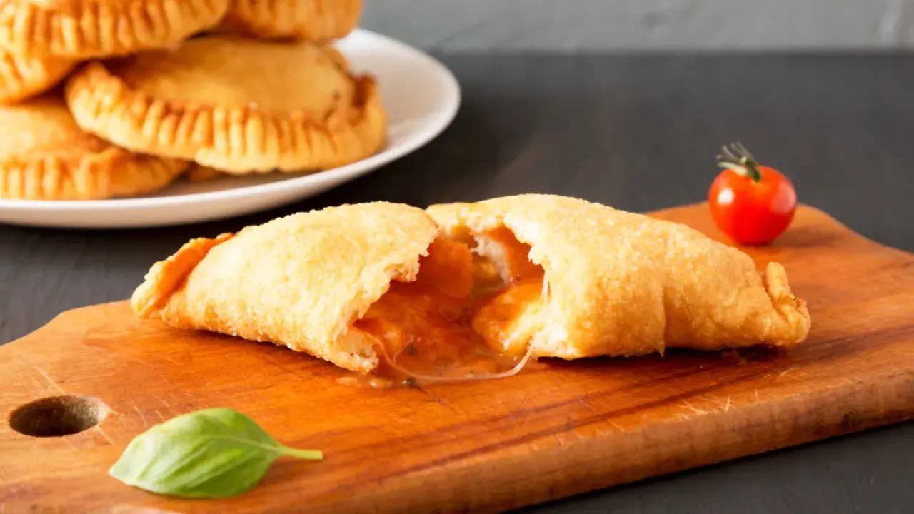 Leckeres Panzerotti-Rezept im Ofen, gefüllt mit Tomaten und Käse. Ein goldbrauner Teig, der auf einer Holzplatte mit Basilikum und Kirschtomate serviert wird.