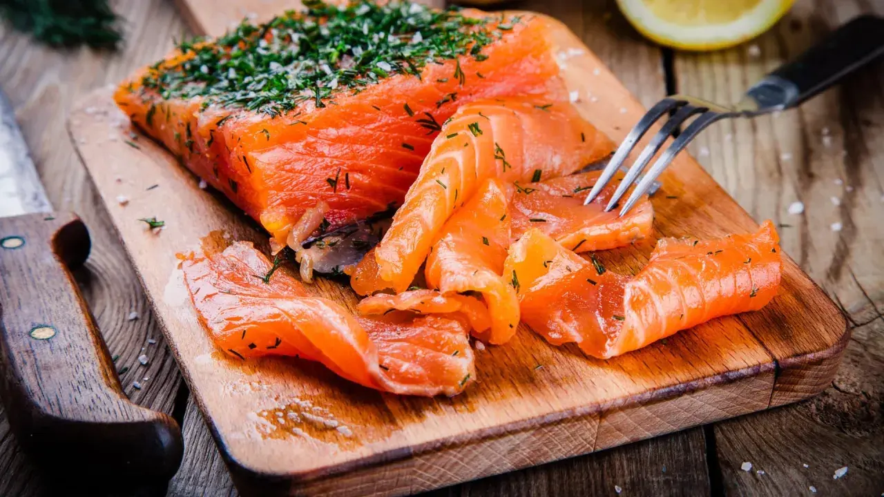 Frisch gebeizter Lachs mit Dill und Salz auf einem Holzbrett. Eine Gabel nimmt ein Stück Lachs auf.