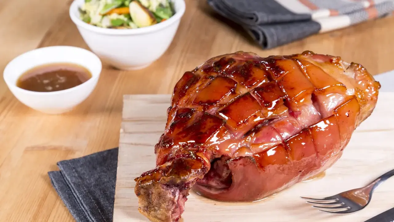 Glänzende Schweinshaxe sous vide auf Holzbrett, daneben Schüssel mit Sauce und Salat.