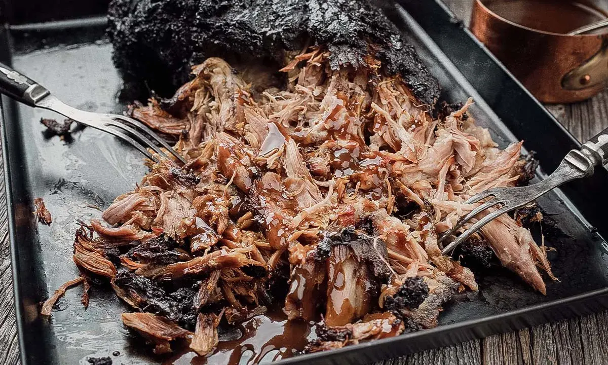 Saftiges Pulled Pork, frisch aus dem Smoker, wird mit Gabeln zerzupft und mit BBQ-Sauce beträufelt.