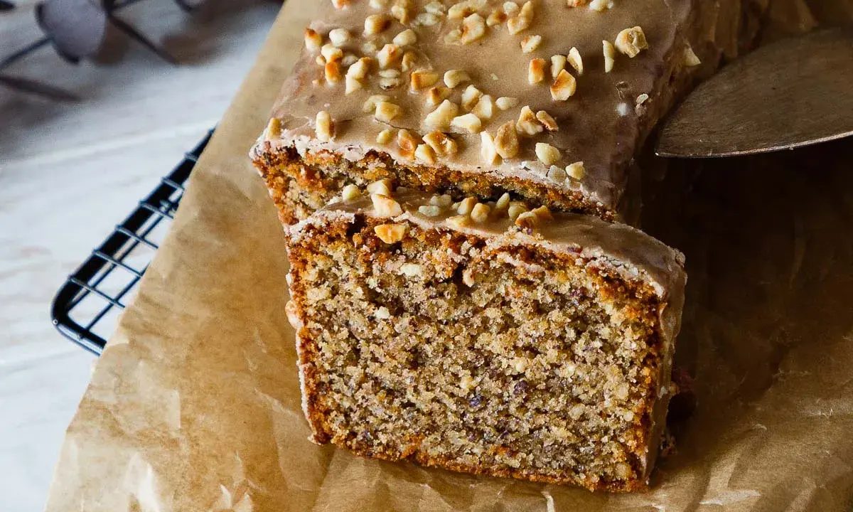 Omas Nusskuchen in Kastenform, mit Zuckerguss und gehackten Nüssen bestreut, wird auf Backpapier präsentiert.