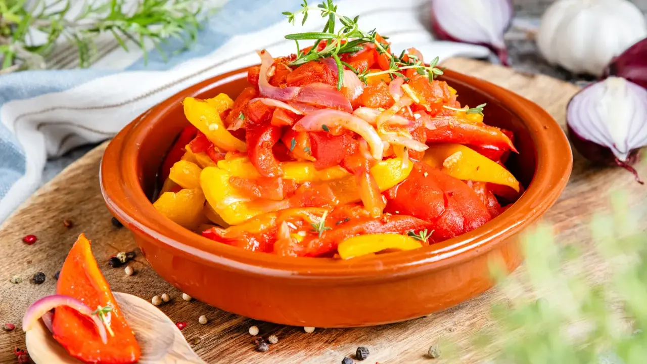 Buntes Peperonata-Gericht im Tontopf, garniert mit Kräutern. Ein Löffel zeigt ein Stück Paprika mit Zwiebeln, perfekt für das original Rezept.