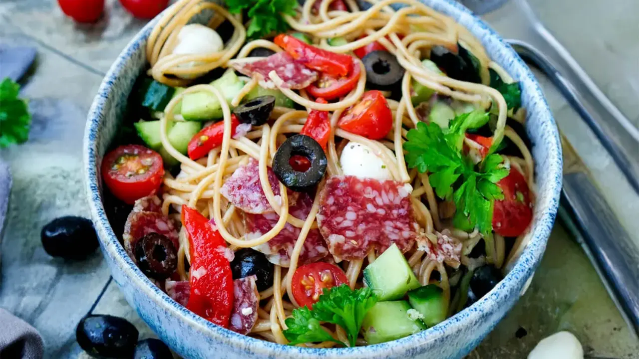 Bunter Spaghetti-Salat mit Salami, Tomaten, Oliven und Mozzarella in einer blauen Schüssel, garniert mit Petersilie.