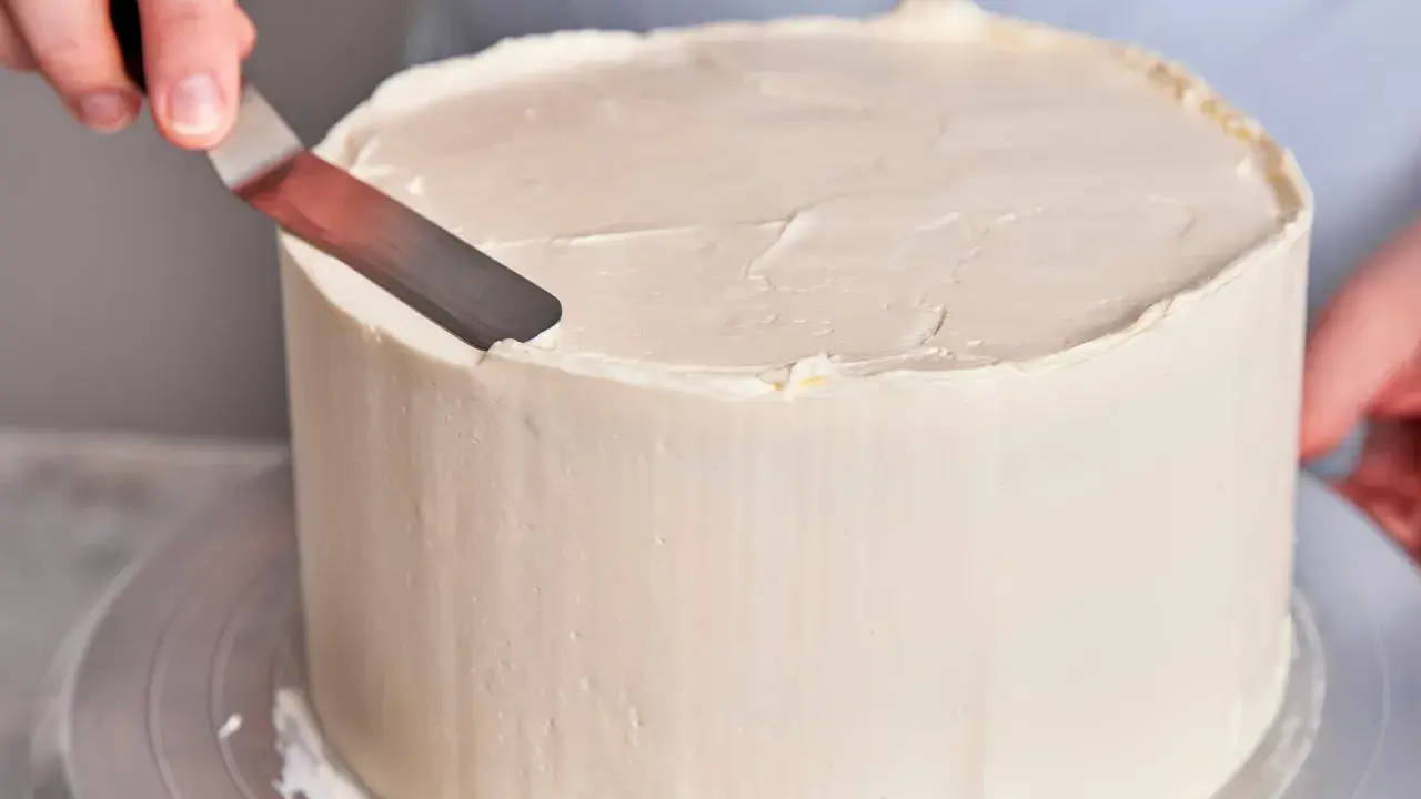 Eine Hand glättet mit einem Spachtel die cremige Buttercreme einer Torte, die für einen Geburtstag bestimmt ist.