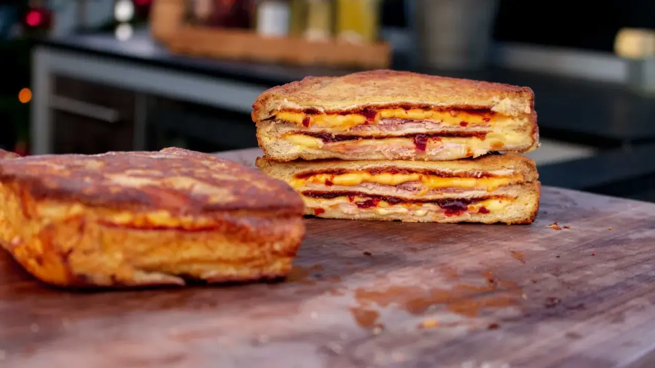 Zwei goldbraune Monte Cristo Sandwiches, gestapelt und mit Käse und Marmelade gefüllt, liegen auf einem Holzbrett.