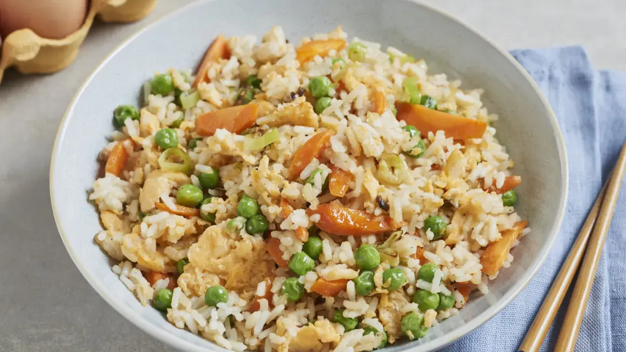 Schüssel mit gebratenem Eierreis wie beim Chinesen, garniert mit Erbsen und Karotten. Daneben liegen Stäbchen.