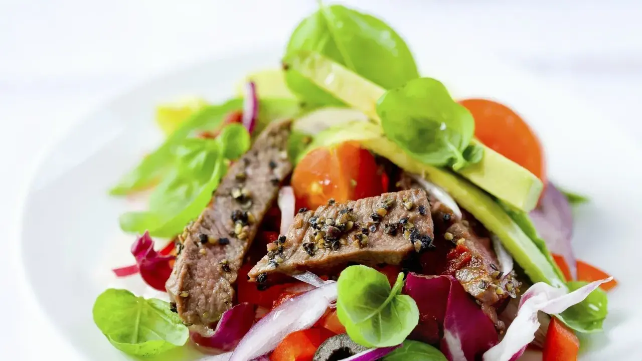 Frischer Salat mit zarten Rinderstreifen, Tomaten, roten Zwiebeln und Basilikum auf einem weißen Teller.