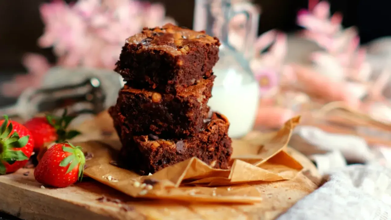 Saftige Brownies nach original Rezept, gestapelt auf einem Holzbrett mit Erdbeeren und einem Krug Milch.