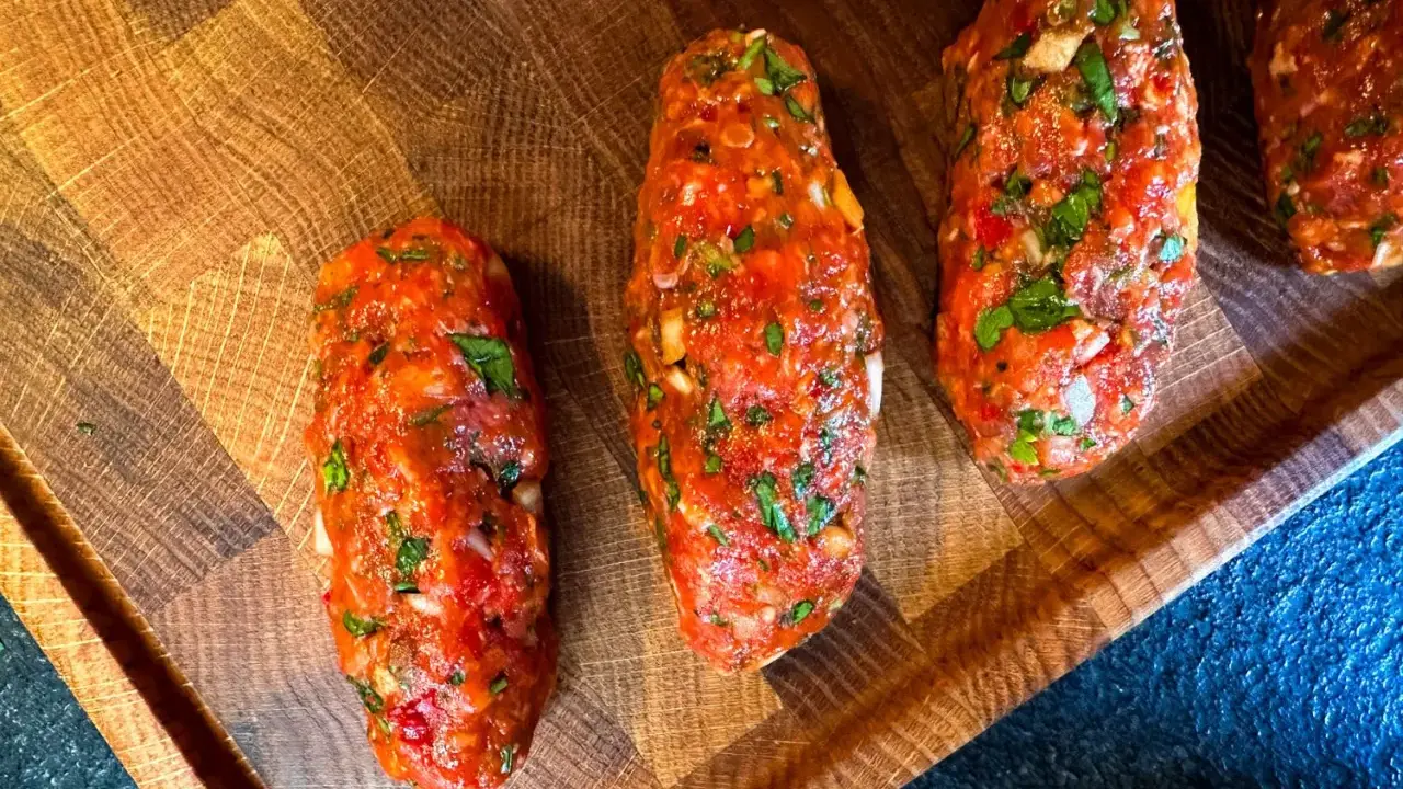 Rohe Cevapcici, bereit für das Rezept, liegen auf einem Holzbrett. Die Hackfleischröllchen sind mit Kräutern und Gewürzen verfeinert.