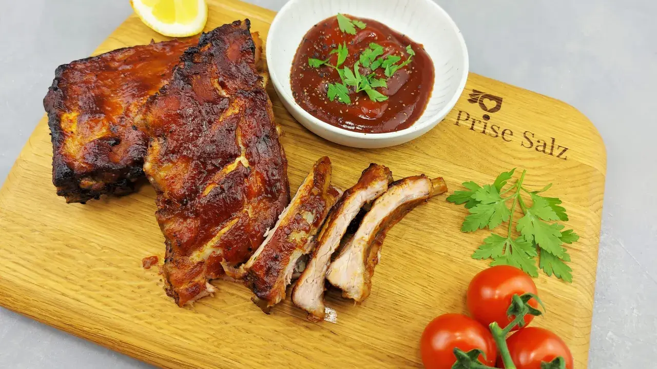 Butterzarte Spareribs im Backofen, glasiert und saftig, serviert mit BBQ-Sauce, Zitrone und frischen Tomaten auf einem Holzbrett.