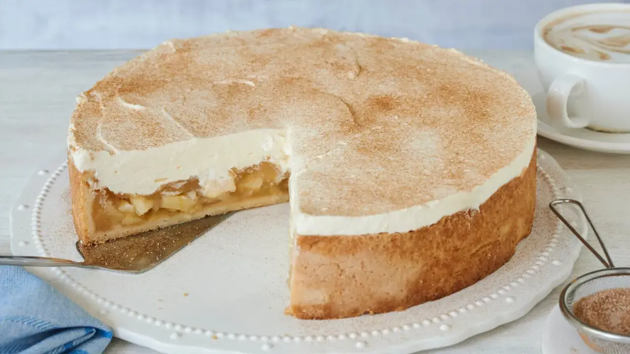 Saftige Apfel-Schmand-Torte mit Zimt bestäubt, ein Stück herausgeschnitten, daneben eine Tasse Cappuccino.