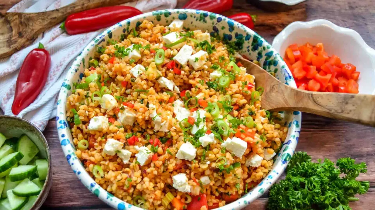 Ein bunter Bulgursalat mit Feta, Paprika und Frühlingszwiebeln, garniert mit Petersilie. Dazu Gurkenscheiben und rote Peperoni.