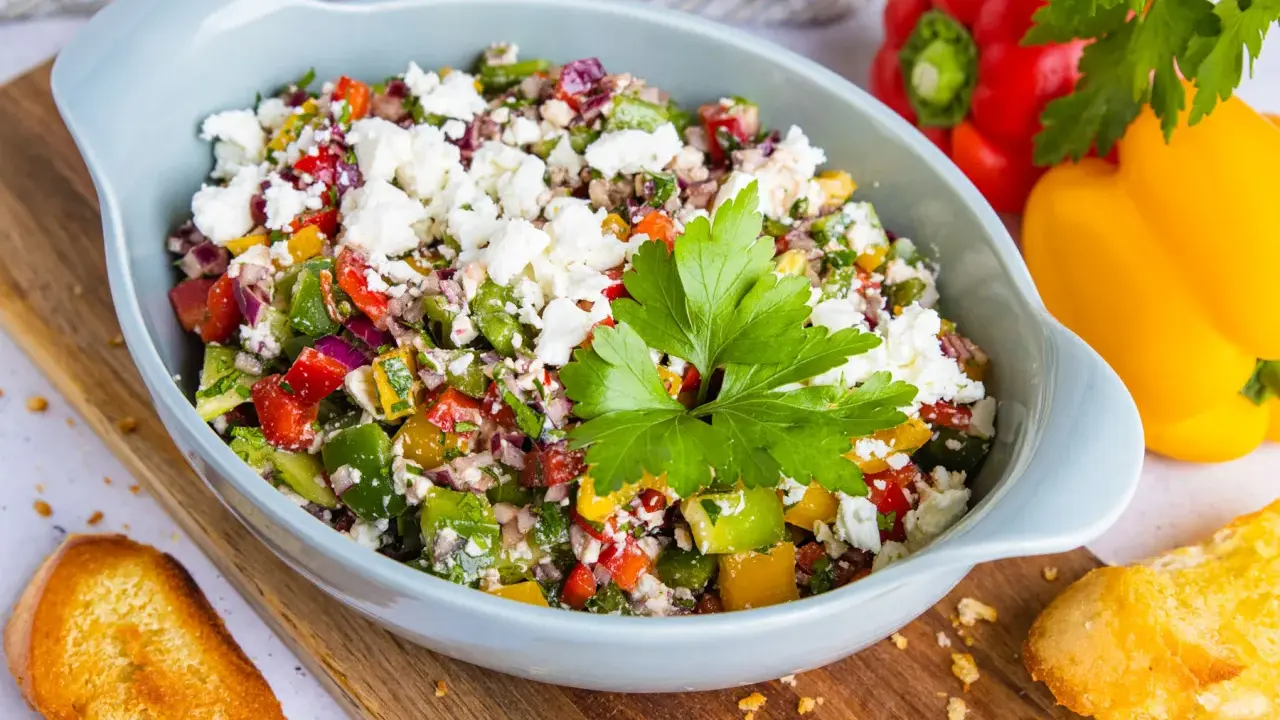 Bunter Paprikasalat mit Feta, garniert mit Petersilie, serviert in einer hellblauen Schüssel auf einem Holzbrett. Daneben liegen geröstete Brotscheiben.