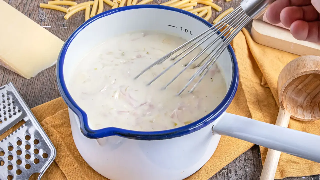 Cremige schinken sahne sauce wird in einem Topf mit einem Schneebesen zubereitet, umgeben von Nudeln, Käse und einer Reibe.