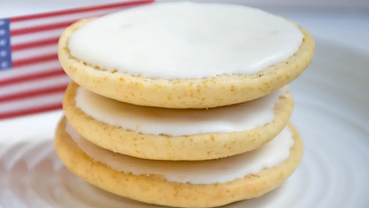 Drei amerikaner Kekse mit weißer Glasur auf einem Teller, im Hintergrund eine kleine US-Flagge. Ein leckeres amerikaner Rezept.
