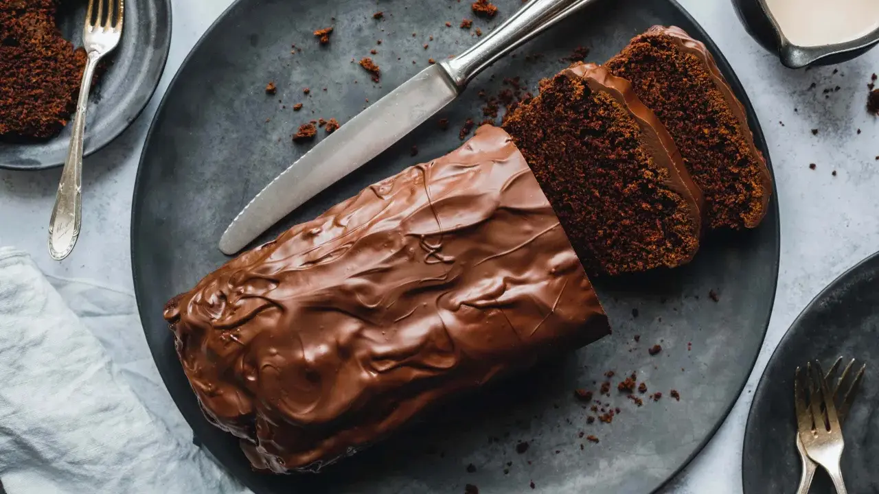 Saftiger Zucchini Schoko Kuchen mit Schokoladenglasur, angeschnitten auf einem dunklen Teller, daneben eine Gabel und ein Messer.