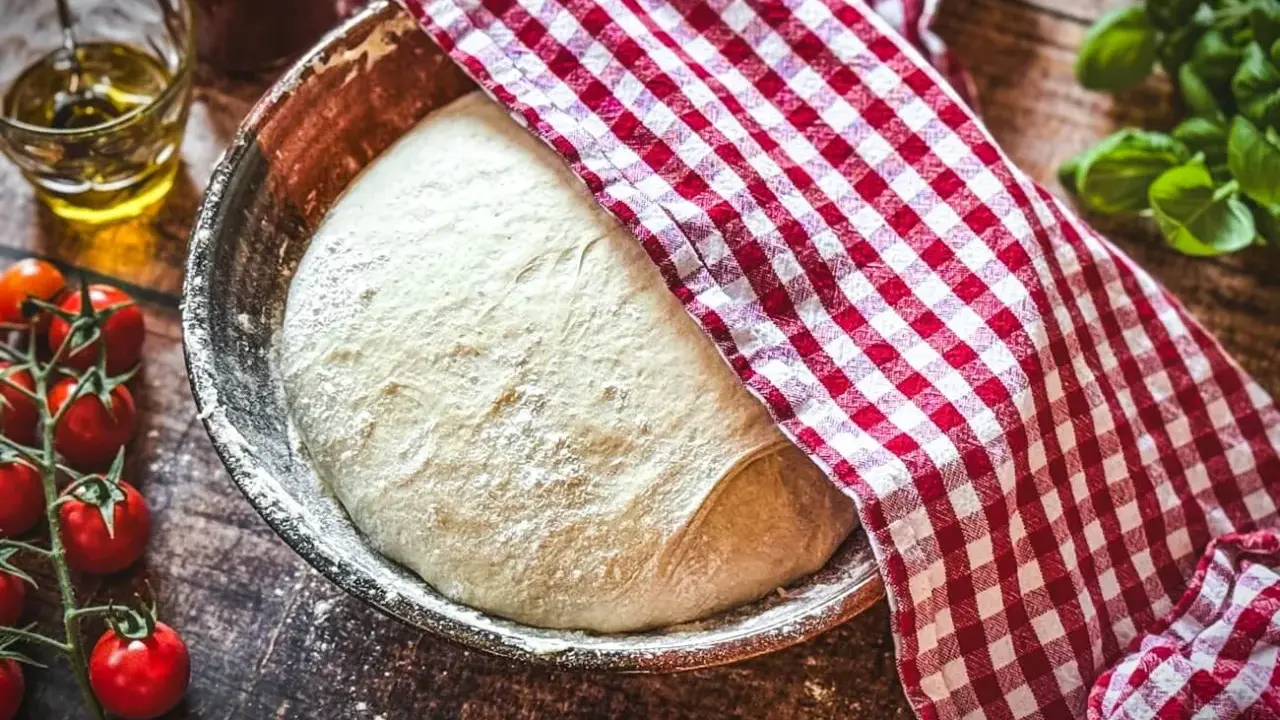 Pizzateig original rezept von einem italienischen gastronomen ruht in einer Schüssel, bedeckt mit einem rot-weißen Tuch. Tomaten und Olivenöl daneben.