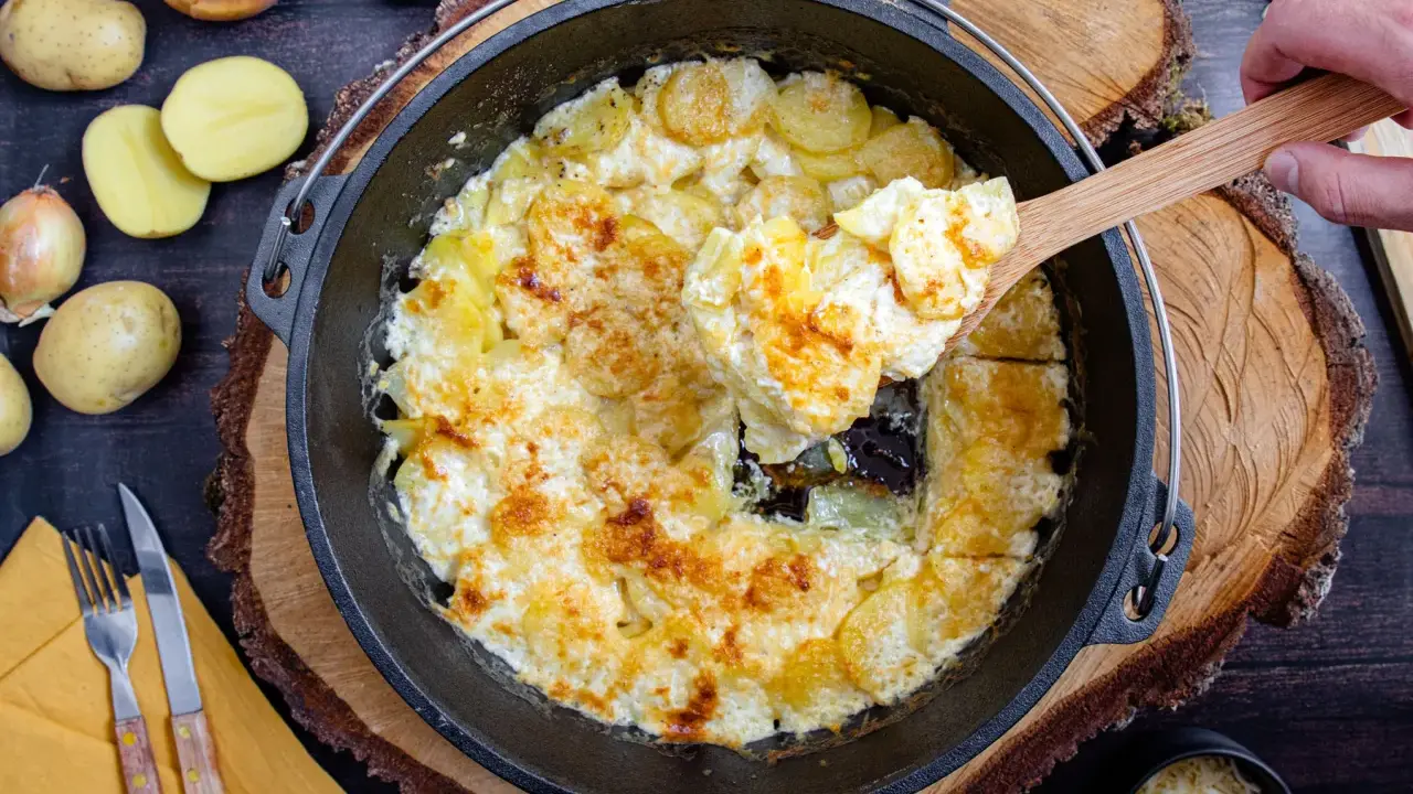 Cremige Kartoffeln im Dutch Oven, goldbraun gebacken. Eine Hand hebt mit einem Holzlöffel eine Portion heraus.