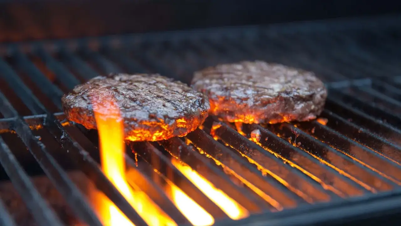 Zwei Burger-Patties auf einem Gasgrill, die Flammen lecken an ihnen hoch. Die richtige Temperatur ist entscheidend für das perfekte Burger-Grillen.