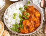 Ein Teller mit dampfendem Reis und cremigem Chicken Tikka Masala, garniert mit frischem Koriander. Dazu Naan-Brot und ein Löffel.
