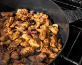 Würziges Gyros brutzelt in einem Dutch Oven auf dem Grill. Zarte Fleischstücke mit Zwiebeln garen in der Pfanne.
