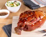 Glänzende Schweinshaxe sous vide auf Holzbrett, daneben Schüssel mit Sauce und Salat.