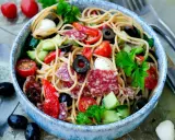 Bunter Spaghetti-Salat mit Salami, Tomaten, Oliven und Mozzarella in einer blauen Schüssel, garniert mit Petersilie.