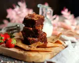 Saftige Brownies nach original Rezept, gestapelt auf einem Holzbrett mit Erdbeeren und einem Krug Milch.
