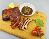 Butterzarte Spareribs im Backofen, glasiert und saftig, serviert mit BBQ-Sauce, Zitrone und frischen Tomaten auf einem Holzbrett.
