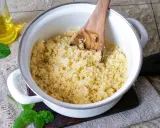 Ein Topf mit lockerem Couscous, bereit für ein marokkanisches Rezept. Ein Holzlöffel rührt die Körner auf. Daneben eine Flasche Öl und frische Minze.