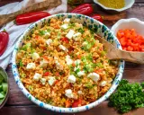 Ein bunter Bulgursalat mit Feta, Paprika und Frühlingszwiebeln, garniert mit Petersilie. Dazu Gurkenscheiben und rote Peperoni.