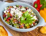 Bunter Paprikasalat mit Feta, garniert mit Petersilie, serviert in einer hellblauen Schüssel auf einem Holzbrett. Daneben liegen geröstete Brotscheiben.