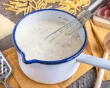 Cremige schinken sahne sauce wird in einem Topf mit einem Schneebesen zubereitet, umgeben von Nudeln, Käse und einer Reibe.