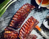 Saftige spareribs grillen holzkohle, mit BBQ-Sauce bestrichen, bereit zum Genuss. Grüne Frühlingszwiebeln und ein Pinsel liegen daneben.