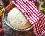 Pizzateig original rezept von einem italienischen gastronomen ruht in einer Schüssel, bedeckt mit einem rot-weißen Tuch. Tomaten und Olivenöl daneben.