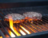 Zwei Burger-Patties auf einem Gasgrill, die Flammen lecken an ihnen hoch. Die richtige Temperatur ist entscheidend für das perfekte Burger-Grillen.