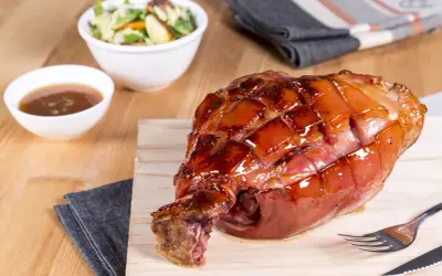 Glänzende Schweinshaxe sous vide auf Holzbrett, daneben Schüssel mit Sauce und Salat.