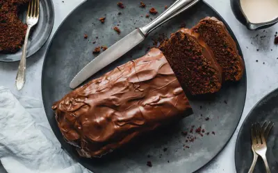 Saftiger Zucchini Schoko Kuchen mit Schokoladenglasur, angeschnitten auf einem dunklen Teller, daneben eine Gabel und ein Messer.