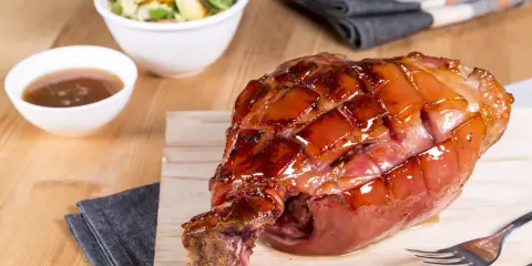 Glänzende Schweinshaxe sous vide auf Holzbrett, daneben Schüssel mit Sauce und Salat.