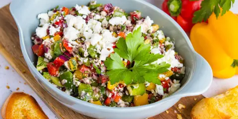 Bunter Paprikasalat mit Feta, garniert mit Petersilie, serviert in einer hellblauen Schüssel auf einem Holzbrett. Daneben liegen geröstete Brotscheiben.
