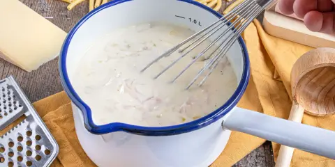 Cremige schinken sahne sauce wird in einem Topf mit einem Schneebesen zubereitet, umgeben von Nudeln, Käse und einer Reibe.