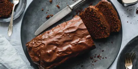 Saftiger Zucchini Schoko Kuchen mit Schokoladenglasur, angeschnitten auf einem dunklen Teller, daneben eine Gabel und ein Messer.