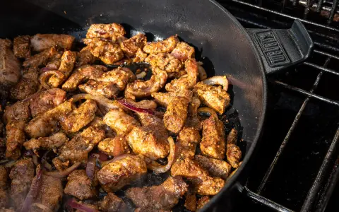 Würziges Gyros brutzelt in einem Dutch Oven auf dem Grill. Zarte Fleischstücke mit Zwiebeln garen in der Pfanne.
