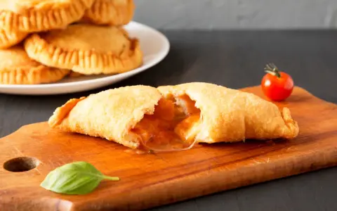Leckeres Panzerotti-Rezept im Ofen, gefüllt mit Tomaten und Käse. Ein goldbrauner Teig, der auf einer Holzplatte mit Basilikum und Kirschtomate serviert wird.