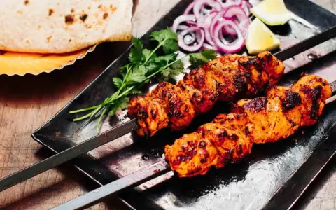 Zwei Spieße mit saftigem Tandoori Chicken, garniert mit Koriander, Zwiebelringen und Zitronenspalten. Dazu Fladenbrot.