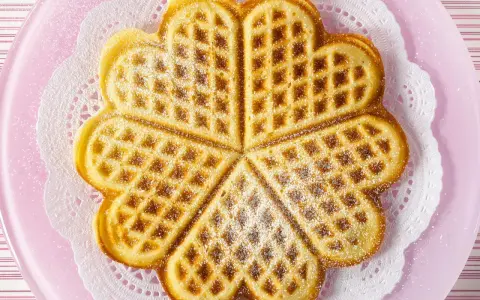 Herzförmige Waffeln ohne Ei, bestäubt mit Puderzucker, auf einem rosa Teller mit Spitzendeckchen.