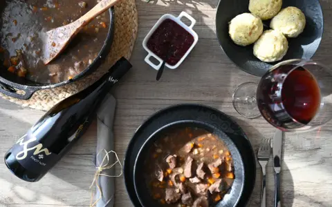 Herzhaftes Wild im Dutch Oven, serviert mit Klößen, Preiselbeeren und einem Glas Rotwein. Ein Festmahl!